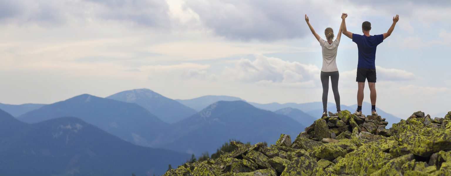 back view of young couple, athletic boy and slim girl standing with raised arms on rocky mountain top enjoying breathtaking summer mountain view. tourism, traveling and healthy lifestyle concept.